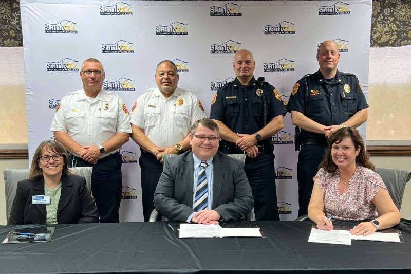 2 male police officials and 2 male fire officials standing behind three people sitting at a table