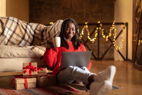 female student in red sweater holding coffee mug sitting on the floor looking at her laptop during the holidays