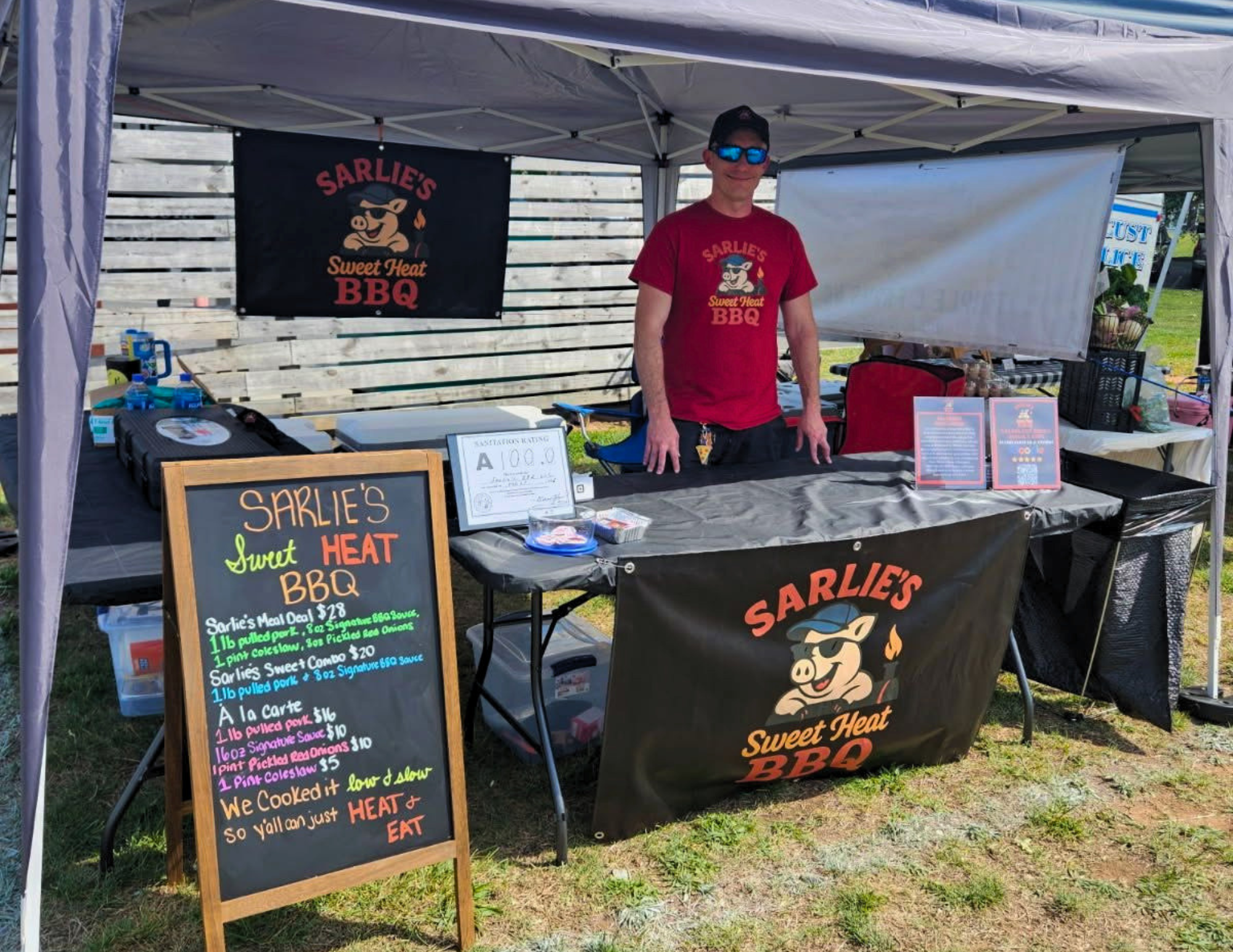Sarlie’s Sweet Heat BBQ owner stands behind table at an outdoor market.